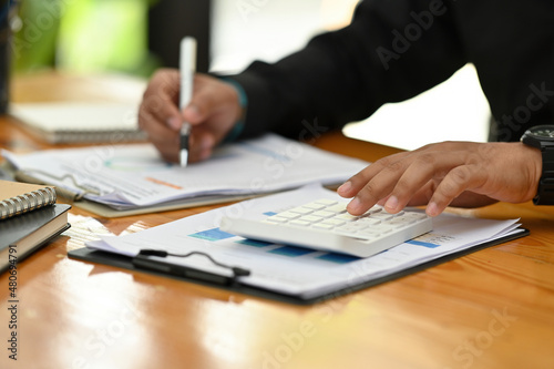 Photo of a young accountant man checking an annual business profit at the wooden table surrounded by a document and calculator.