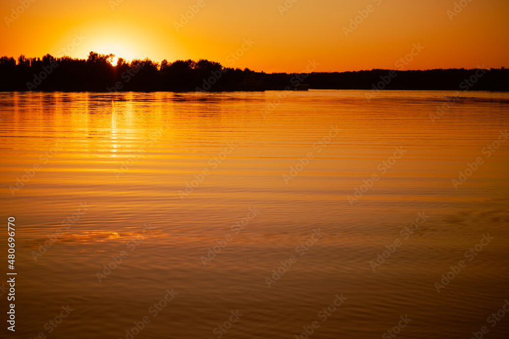 Shining lake with sun setting below horizon and covering with rays water surface with dark trees in background. Astonishing view full of yellow and orange colors. Shining lake with sun setting below horizon and covering with rays water surface with dark trees in background. Astonishing view full of yellow and orange colors.