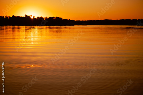 Shining lake with sun setting below horizon and covering with rays water surface with dark trees in background. Astonishing view full of yellow and orange colors.