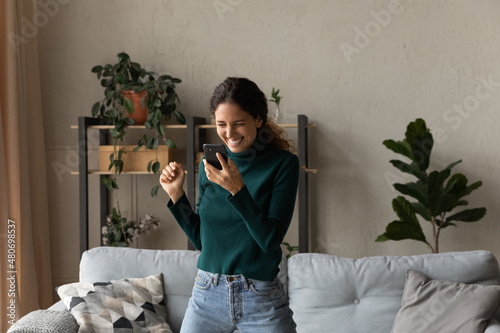 Photos Happy euphoric millennial generation hispanic latin woman looking at mobile phone screen, feeling excited reading message with amazing win news, jumping dancing alone in living room, internet success