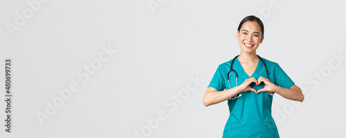 Covid-19, healthcare workers, pandemic concept. Lovely caring asian doctor, female nurse in scrubs showing heart gesture and smiling, taking care of patients with love, white background