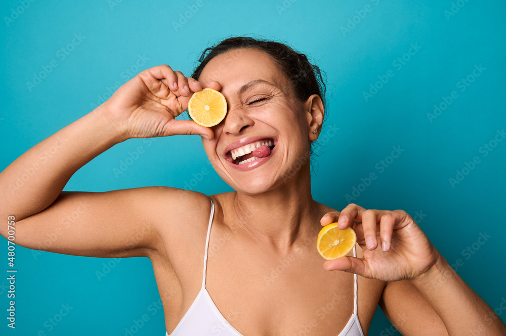 Attractive delighted young woman smiling with cheerful beautiful healthy toothy smile, holding lemon halves and covering her eyes with it. Isolated on bright blue background with copy ad space Attractive delighted young woman smiling with cheerful beautiful healthy toothy smile, holding lemon halves and covering her eyes with it. Isolated on bright blue background with copy ad space