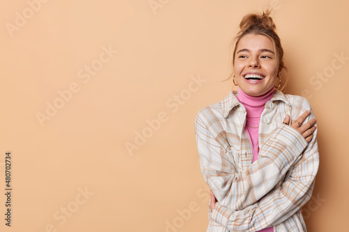 Studio shot of happy carefree woman looks gladfully away feels cheerful dressed in casual comfortable soft clothes poses against beige studio wall with copy space for your promotional content