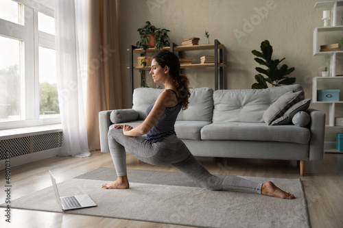 Full length happy active young hispanic latina woman in sportswear stretching watching online lesson on computer  enjoying webcam distant class with trainer  improving flexibility in living room.