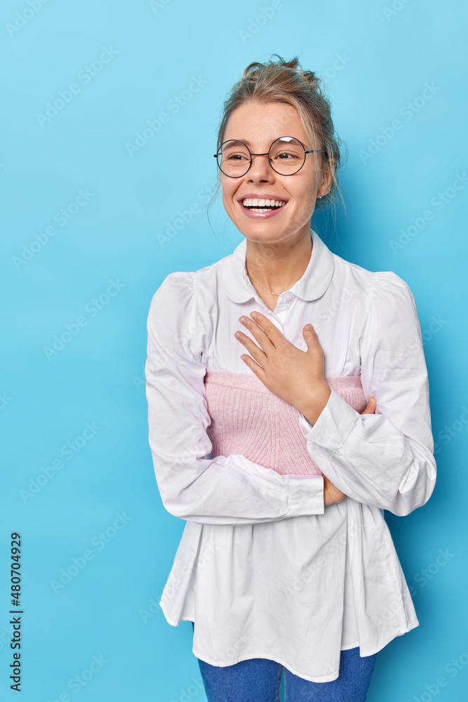 Positive young businesswoman feels glad after successful formal meeting keeps hand on chest and smiles happily dressed in formal outfit poses against blue studio background. Happy office worker Positive young businesswoman feels glad after successful formal meeting keeps hand on chest and smiles happily dressed in formal outfit poses against blue studio background. Happy office worker
