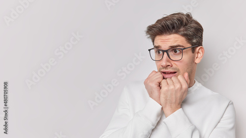 Stressed worried man keeps hands on chin trembles from fear concentrated away feels embarraassed wears transparent glasses and jumper isolated over white background blank space on left side.