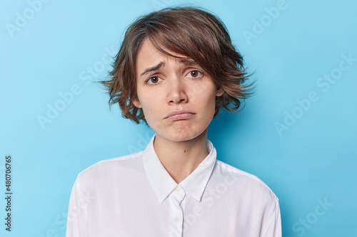 Portrait of frustarted young woman feels discontent frowns face has bad mood dressed in elegant white shirt feels disappointed isolated over blue background. Negative human emotions and feelings