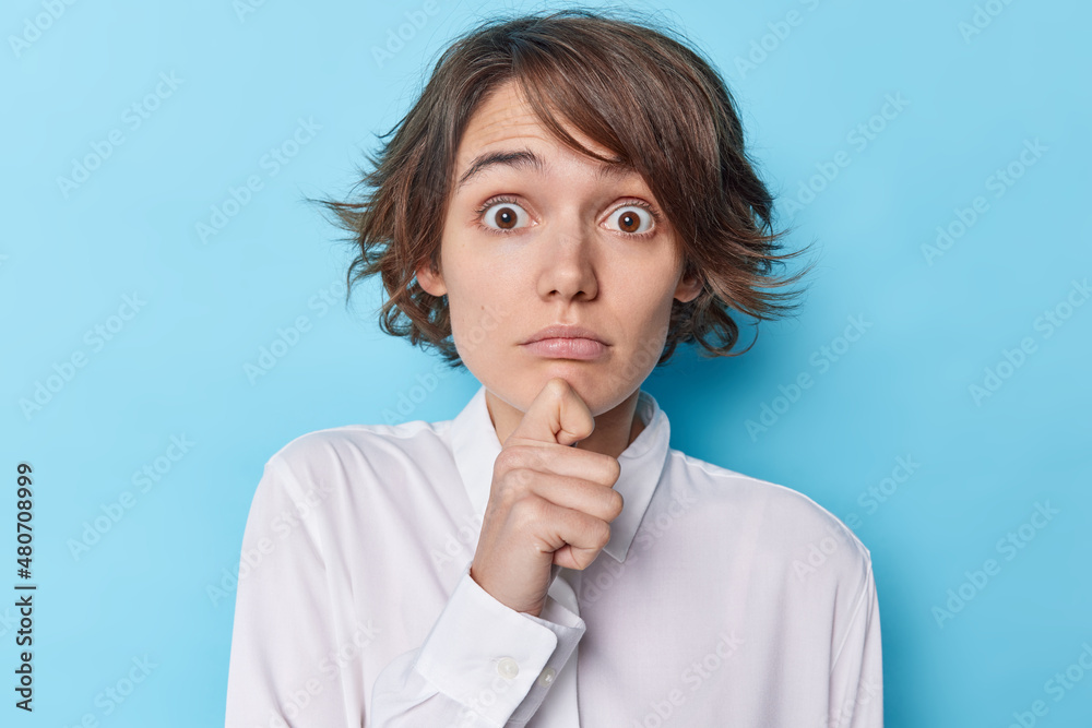 Portrait of impressed young woman holds chin stares with popped eyes hears shocking news has trendy hairstyle wears white shirt isolated over blue background stands overwhelmed. Human reaction Portrait of impressed young woman holds chin stares with popped eyes hears shocking news has trendy hairstyle wears white shirt isolated over blue background stands overwhelmed. Human reaction