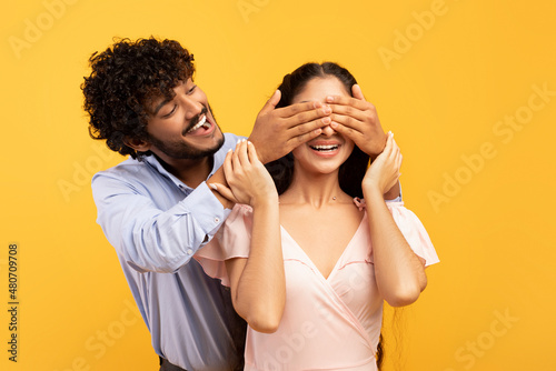 Romantic surprise concept. Portrait of happy indian guy covering his pretty girlfriend eyes from back, yellow background