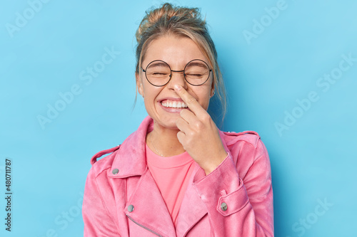 Cheerful woman with combed hair touches nose foolishes around wears spectacles and pink jacket isolated over blue background tries to amuse someone. Positive fair haired girl has fun indoor.