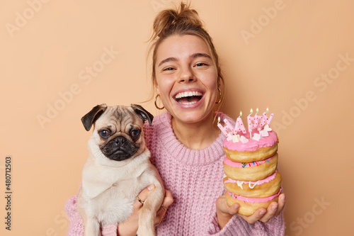 Cheerful positive woma has festive mood celebrates birthday of her pug dog holds delicious doughnuts with burning candles invites you to join celebration wears knitted sweater isolated over beige wall photo