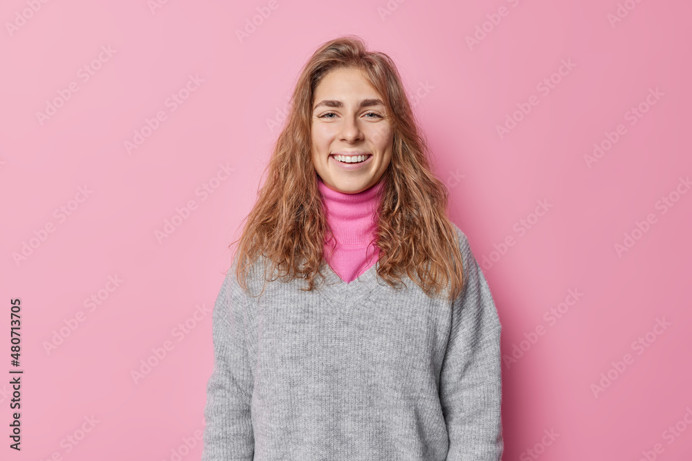 Fototapeta premium Beautiful happy woman with long hair smiles gladfully has clean natural skin ni make up feels happy wears casual jumper poses against pink background. Positive human face expressions and feelings