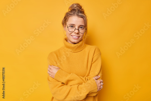 Young attractive woman with hair bun embraces herself smiles gently enjoys softness of new jumper looks gently at camera isolated over yellow background. Positive human facial expressions concept © Wayhome Studio