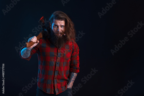 Emotional man with cigar holds old axe. Bearded lumberjack in checkered shirt with long hair
