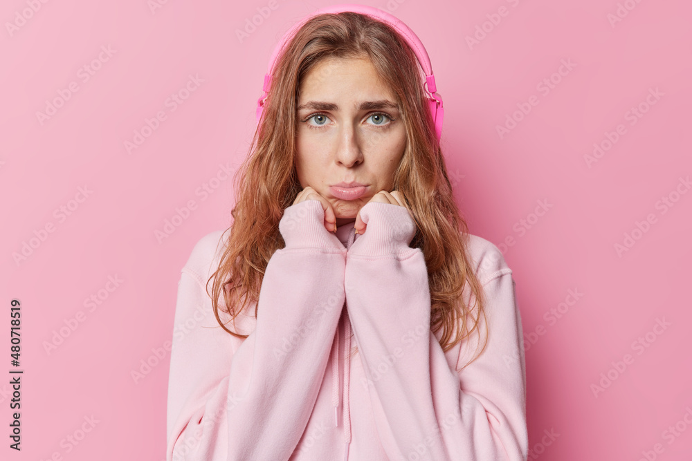 Unhappy displeased beautiful woman with blue eyes purses lis keeps hands under chin feels frustrated has bad mood listens music via headphones wears casual hoodie isolated over pink background Unhappy displeased beautiful woman with blue eyes purses lis keeps hands under chin feels frustrated has bad mood listens music via headphones wears casual hoodie isolated over pink background