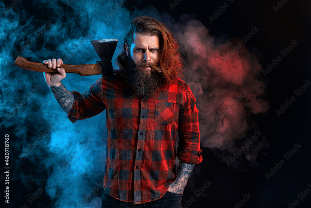Bearded lumberjack. Bearded man with cigar in checkered shirt with long hair holds old axe in smoke