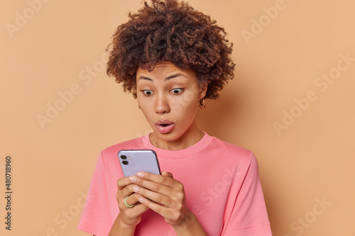 Impressed curly haired woman gets shocking message stares at smartphone keeps mouth widely opened dressed in casual t shirt isolated over beige background finds out surprising news stands indoor