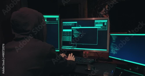 Hacker in a hood sits in front of computer screens and hacks databases. The concept of cyberterrorism, server hacking and cyberattacks. The room is dark, a person is typing code into the command line