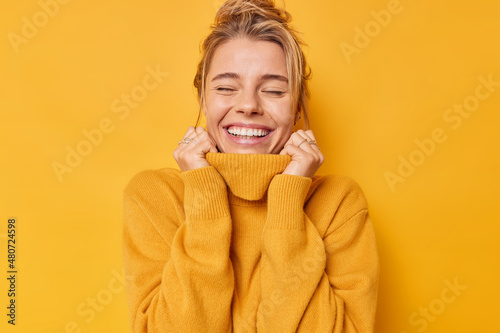 Happy optimistic woman keeps eyes closed smiles gently daydreams holds hands on collar of jumper recalls nice memories isolated over vivid yellow background breathes free. Carefree emotions.