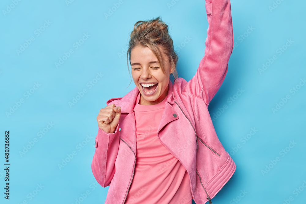 Upbeat European woman shakes arms celebrates victory makes triumph dance exclaims loudly closes eyes wears fashionable pink jacket isolated over blue background achieves goal or wis challenge - obrazy, fototapety, plakaty 