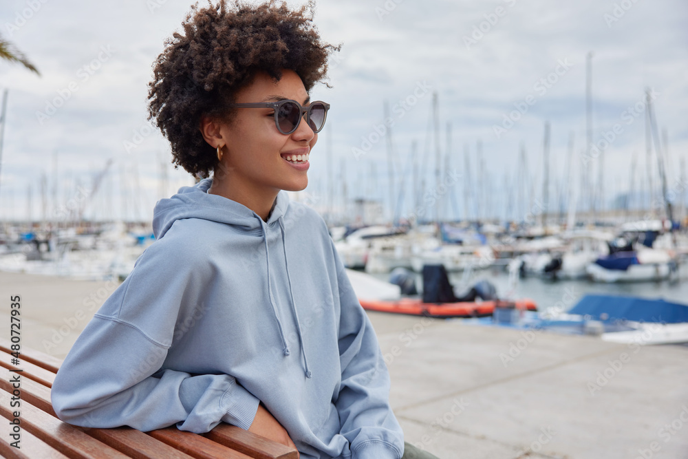 Sideways shot of positive curly haired woman poses on bench wears sunglasses and casual blue hoodie smiles broadly enjoys beautiful view of harbor breathes fresh marine air rests after walking Sideways shot of positive curly haired woman poses on bench wears sunglasses and casual blue hoodie smiles broadly enjoys beautiful view of harbor breathes fresh marine air rests after walking