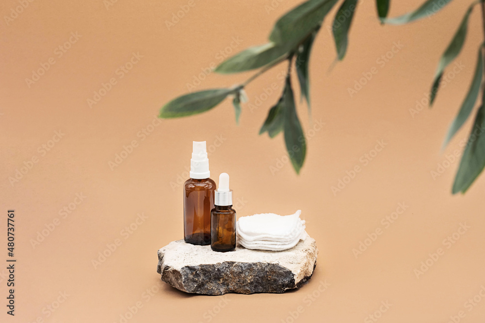Blank amber glass essential oil bottle with pipette, glass spray bottle and reusable cotton sponges on natural stone podium. Organic spa cosmetic beauty product mock up.