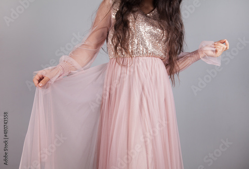 Stylish young woman with pink dress, holding a skirt, on gray background.