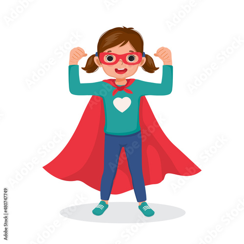 Cute little girl playing dressed in superhero costumes standing raising hands showing her strong arm fists