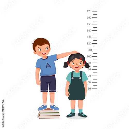 cute little boy standing on stack of books measuring height of little girl growth with measurement ruler on the background of wall