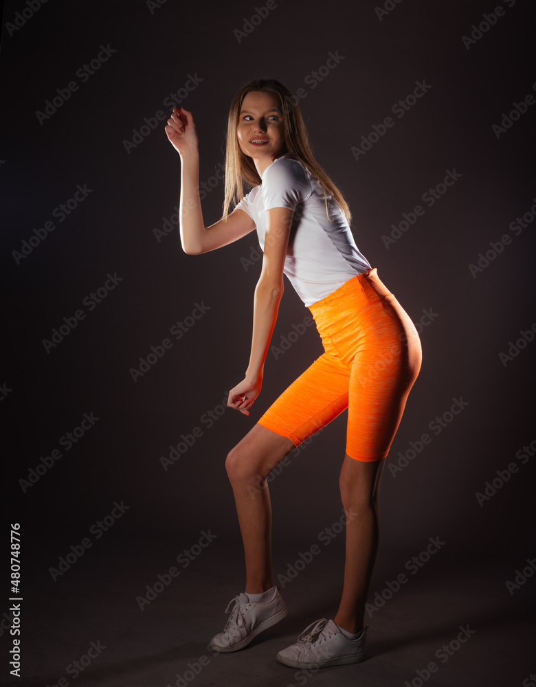 Portrait of a smiling young sportswoman standing in a studio and wearing a white top and short orange leggings Portrait of a smiling young sportswoman standing in a studio and wearing a white top and short orange leggings