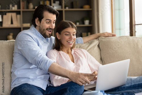 Surprised young couple sit on sofa looking at laptop screen read great news, feels amazed by unbelievable huge discounts, get commercial sale offer, unexpected money income, online lottery win concept