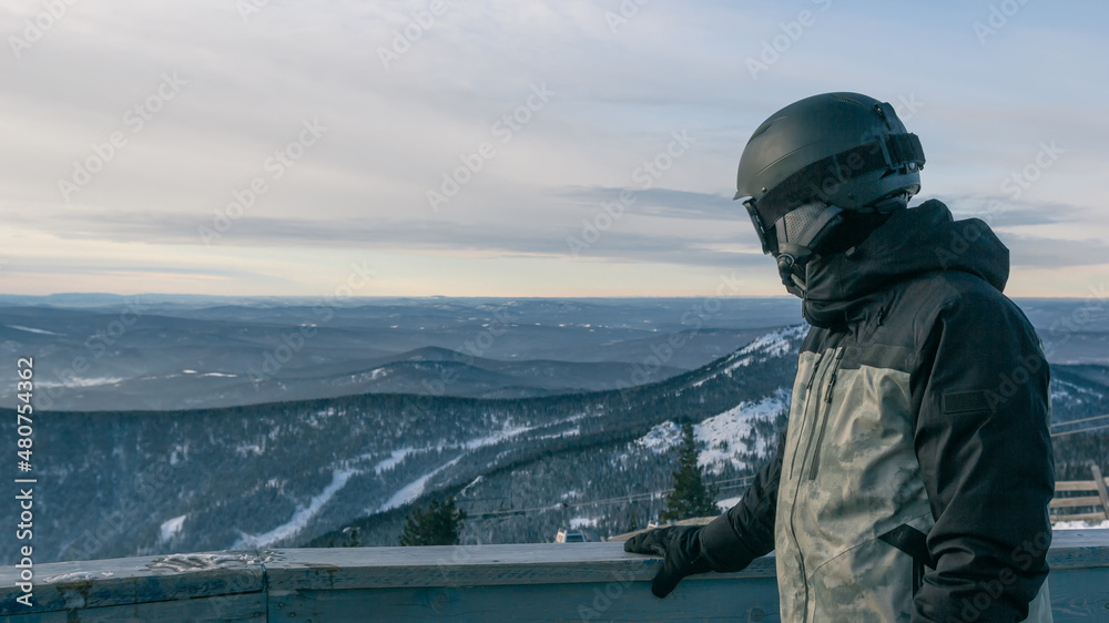 A snowboarder at an observation point in a ski resort leans on the railing and looks at a beautiful winter mountain landscape