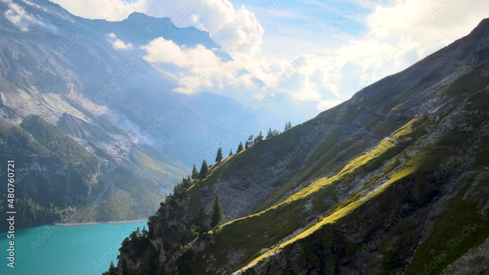 Aerial drone shot of the side of a rocky mountain on a sunny day. Walking path in the middle of the swiss alps on a green cliff with Blue glacier lake Oeschinen in the background