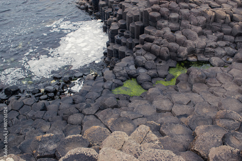 Hexagonal shaped rocks on coast with dark blue wavy water and green seaweed