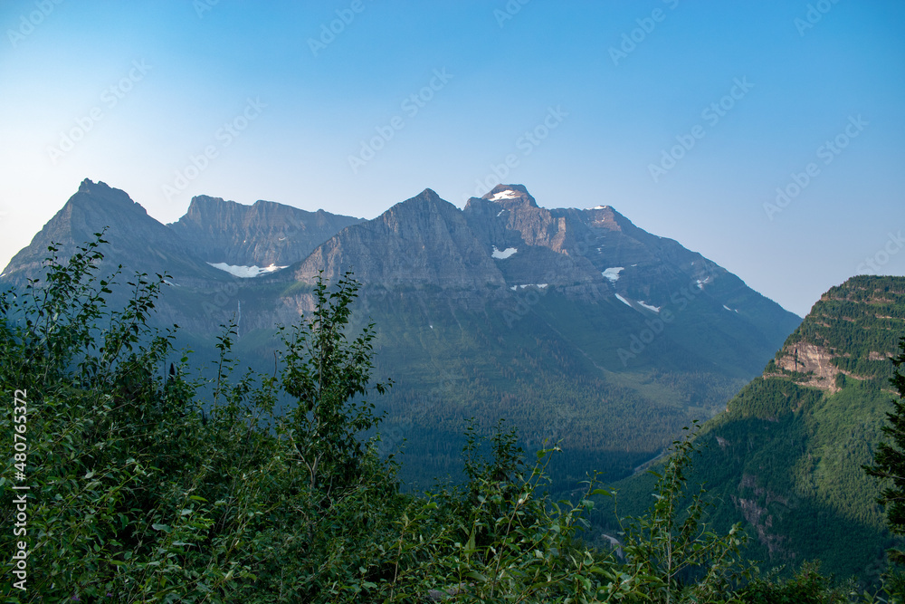 Snowy and rocky mountains seen from forest of green trees with blue sky in Glacier National Park
