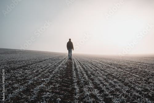 Young caucasian man in jacket standing on meadow, field covered with hoarfrost in misty fog at sunrise. Czech landscape