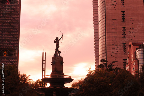 cdmx, distrito fedral de mexico/mexico; 1,15,2020: fountain of the hunting target at sunset (fuente de la diana cazadora) . orange clouds can be seen in the middle of two buildings in the background.
