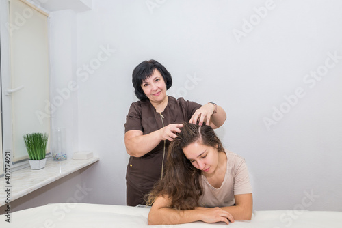 A female masseuse makes a head massage to a young beautiful girl who is sitting at the massage table.