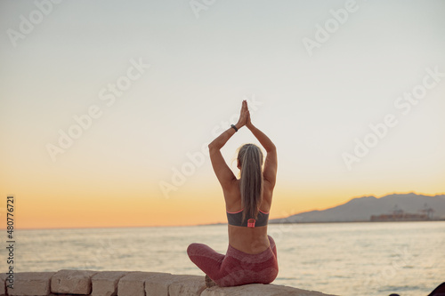 Back view of healthy female practicing yoga, sitting on edge at seaside on the background of sky at sunset