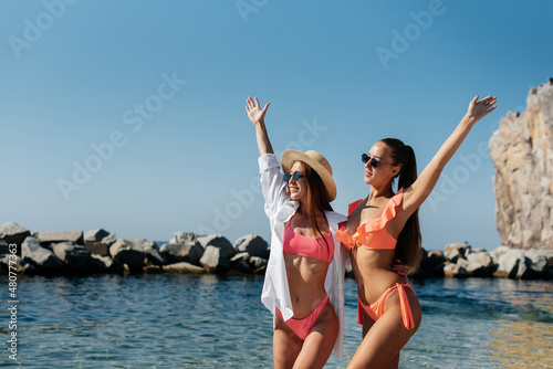 Two beautiful young girls in glasses, bikinis and hats are standing and enjoying on the ocean shore against the backdrop of huge rocks on a sunny day. Tourism and tourist trips.