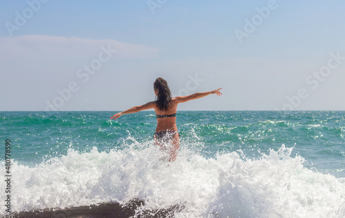 A girl in a swimsuit looks into the sea, enjoys the splashes of the wave. Beautiful waves. Rest by the sea. The sea season.