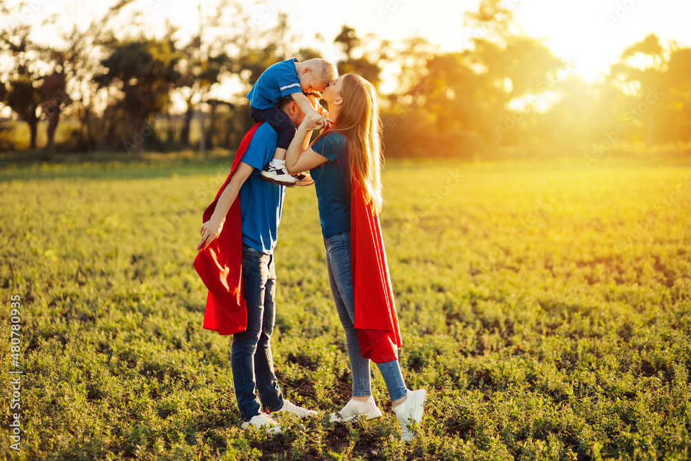 Mom Dad and child play together in the park. Parents and son in Superhero costumes cheerfully smiling and hugging