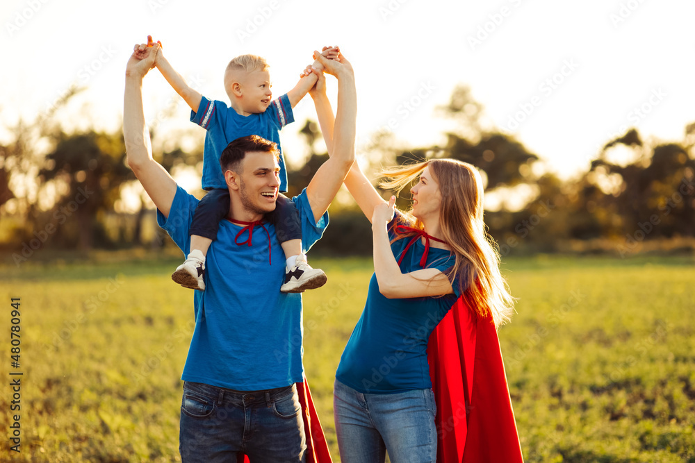 Fototapeta premium Mother, father and son in superhero costumes. Parents hold their son in their arms and play superhero outdoors