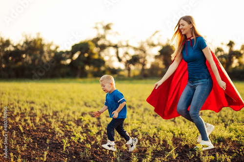 Obraz na płótnie Super mom and her son are running forward. Cheerful family, woman in a red cape as a superhero w salonie