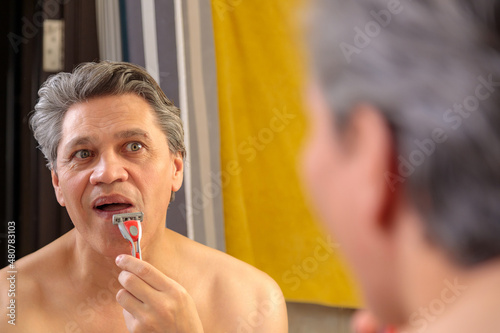 An adult gray-haired man shaves with a razor in front of a mirror