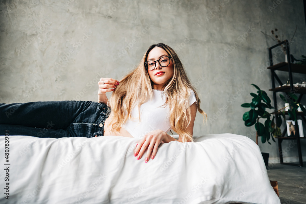 Fototapeta premium Young attractive caucasian woman in eyeglasses with blonde hair sitting on a bed home.