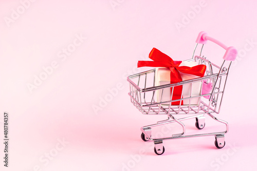Bright pink shopping trolley push cart with white gift box on light rose background with copy space. Valentines day celebration sale, romantic present discounts concept.