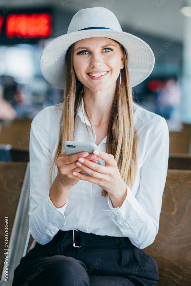 Millennial woman texting friends or writing email on smartphone while sitting with baggage in modern airport lounge, businesswoman go on business or work trip waiting for flight.