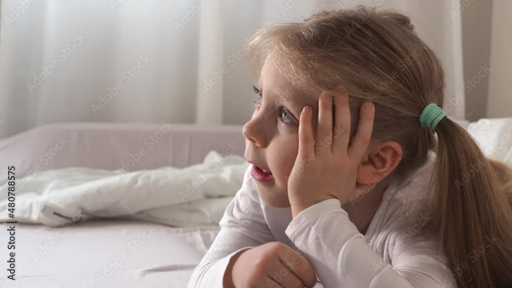 A beautiful little preschooler girl in white pajamas on the bed watches TV intently. The concept of children's addiction. Watch over the children. Day off, hobby. Children and TV Addiction A beautiful little preschooler girl in white pajamas on the bed watches TV intently. The concept of children's addiction. Watch over the children. Day off, hobby. Children and TV Addiction