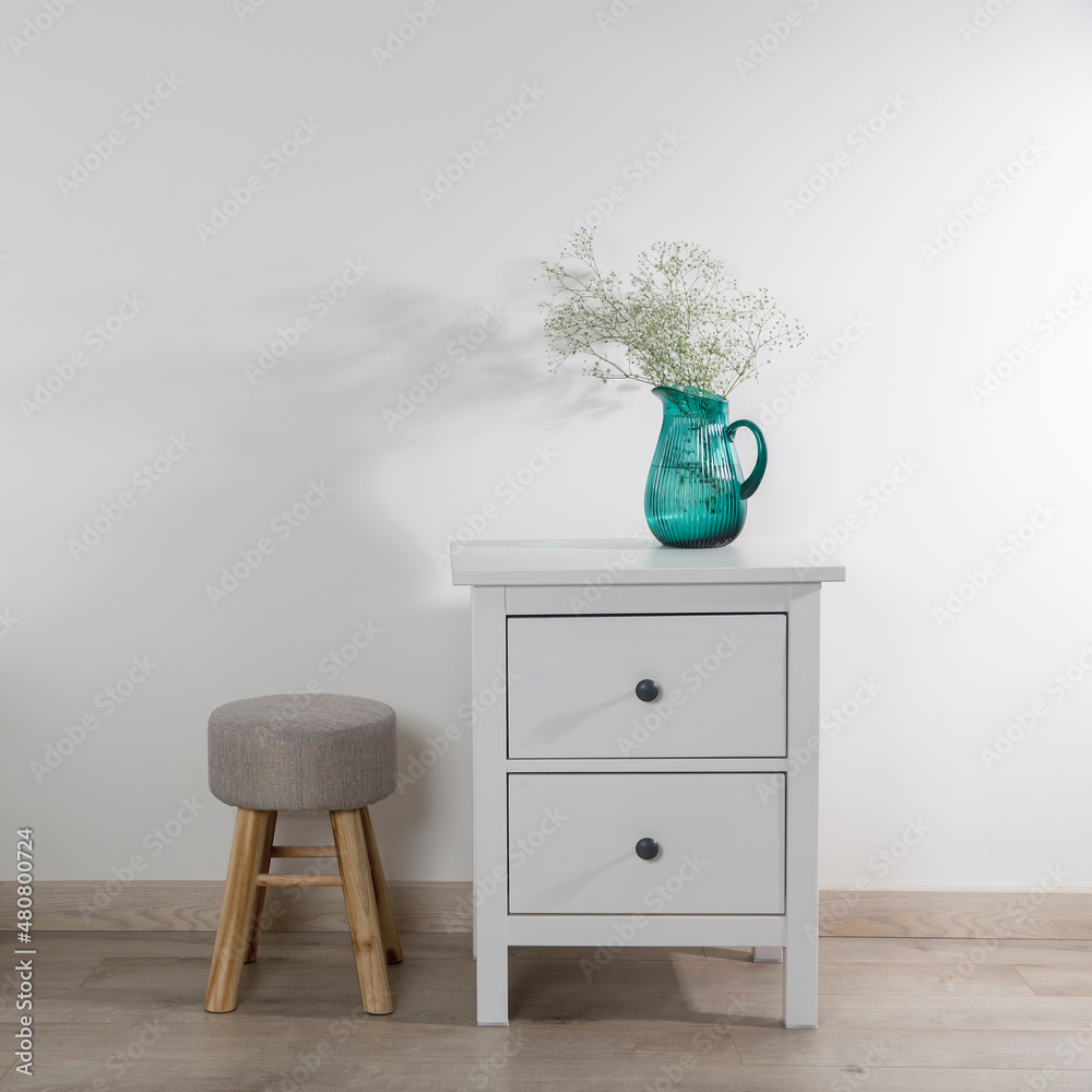 Φωτογραφία A large glass green jug with a bouquet of gypsophila, a white cup, a teapot and a notepad on a chest of drawers, a stool and a wicker laundry basket